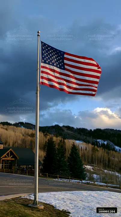 American flag flying in strong wind above a landscape with mountains and trees.
