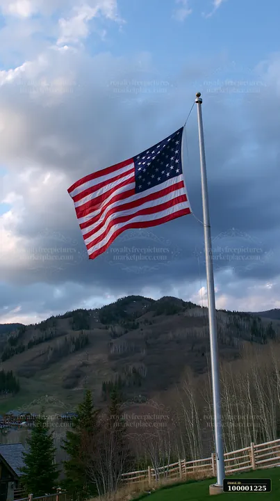 An American flag flying in strong wind stands tall on a mountaintop surrounded by trees and hills.