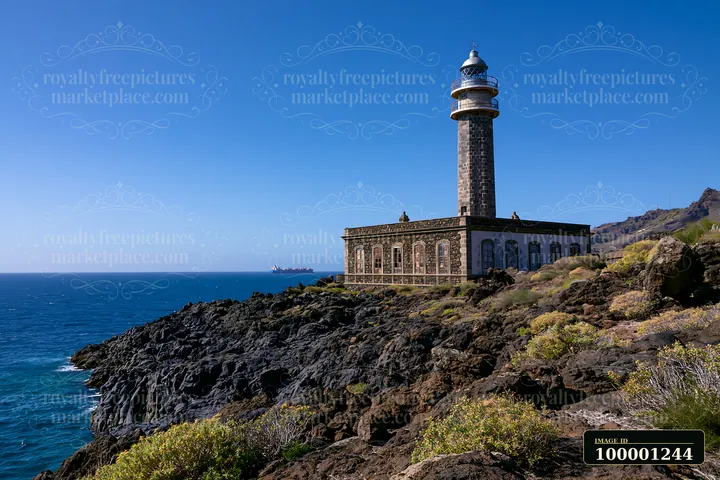 A lighthouse stands on rocky land with a cargo ship visible on the horizon.