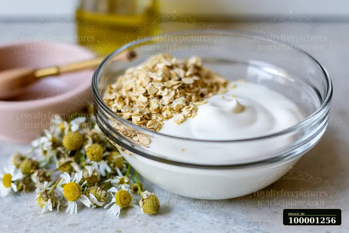 A bowl holds a cream dose with natural skincare ingredients, surrounded by oats and flowers on a countertop.