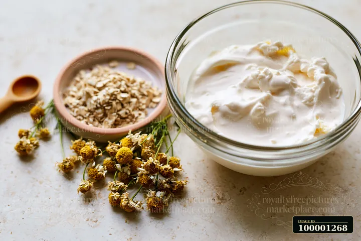 A bowl contains a creamy mixture made with natural ingredients, including oats and dried flowers, arranged on a kitchen surface.