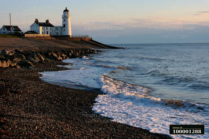 A lighthouse stands on the shore as waves wash over a pebbly beach during sunset.
