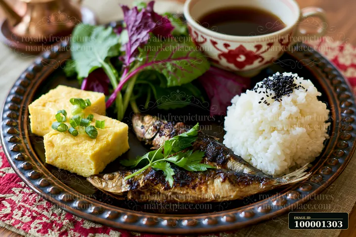 A breakfast plate with Japanese breakfast includes grilled fish, rice, rolled omelette, and mixed greens, arranged on a decorative tray with tea.