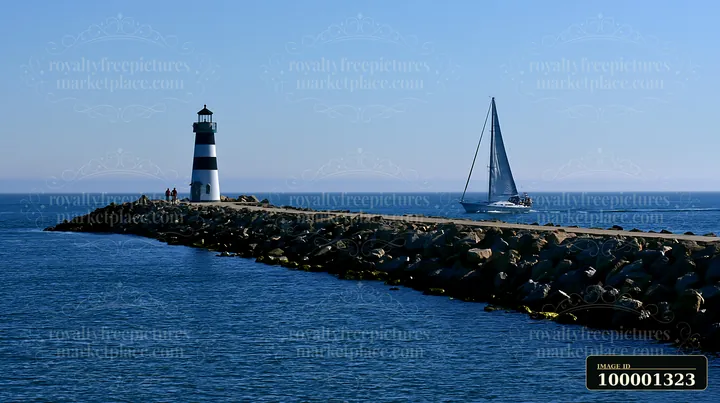 A lighthouse with black and white stripes stands at the end of a jetty while a sailboat approaches it under a clear blue sky.