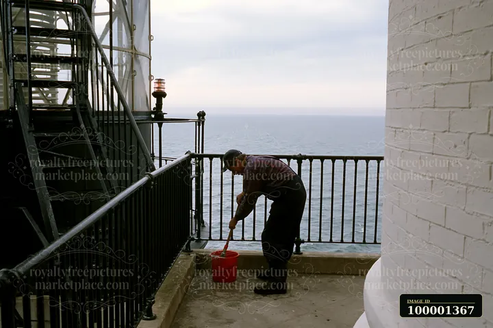 A lighthouse maintenance worker is cleaning the area around the lighthouse, using a bucket and brush.