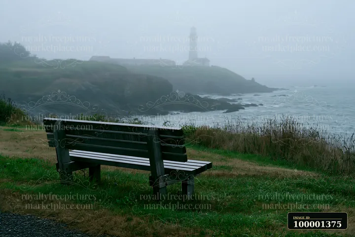 A lighthouse in heavy ocean fog stands on a rocky shore with a bench in the foreground.
