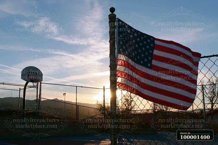 An American flag waving in gentle breeze stands near a basketball court at sunset.