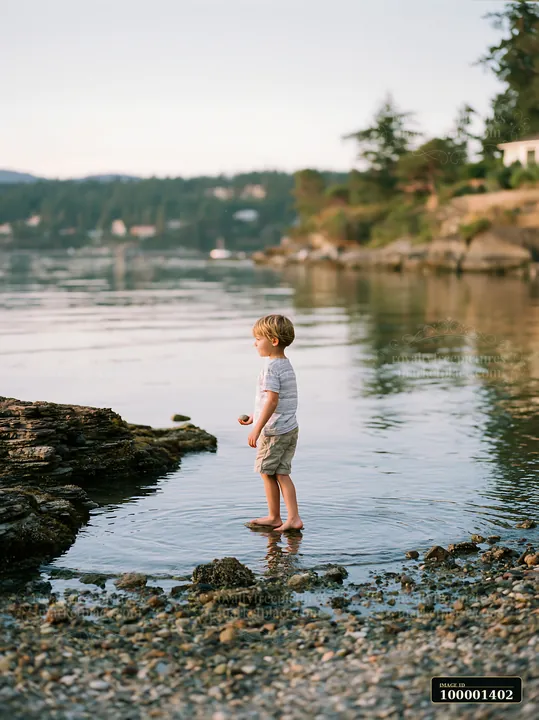 A young boy stands in shallow water at a coastline with exposed rocks, holding a small stone.