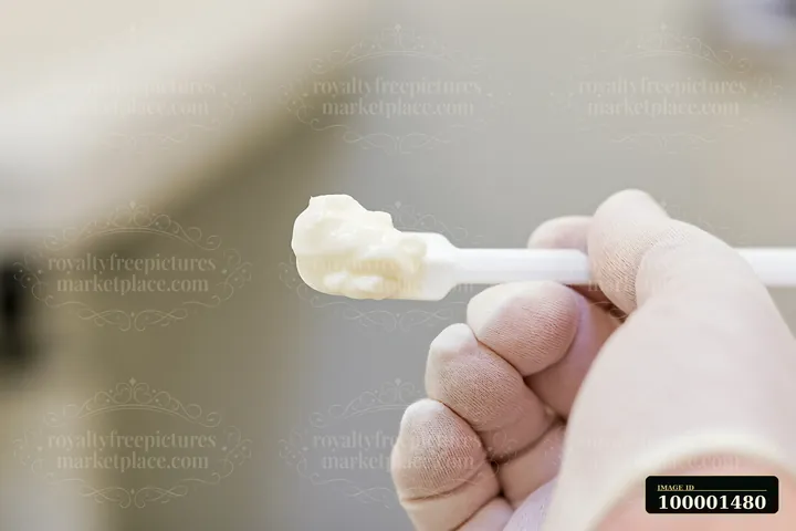 A hand wearing a glove holds a cosmetic spatula with cream dose on a cosmetic spatula in a skincare setting.