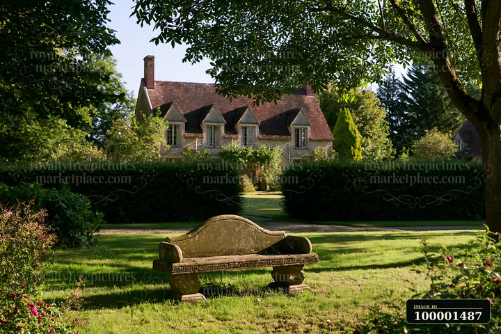 A country house surrounded by hedges sits in a lush garden with a stone bench in the foreground.