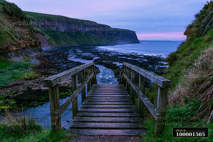 A wooden path leads to the coastline beneath dramatic cliffs, where the waves break against the rocky shore during twilight.