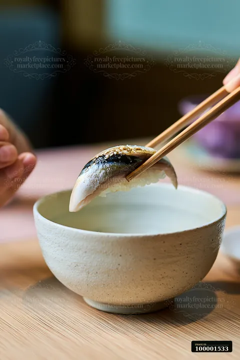 A hand holds a single sushi piece ready to eat with chopsticks above a simple bowl.