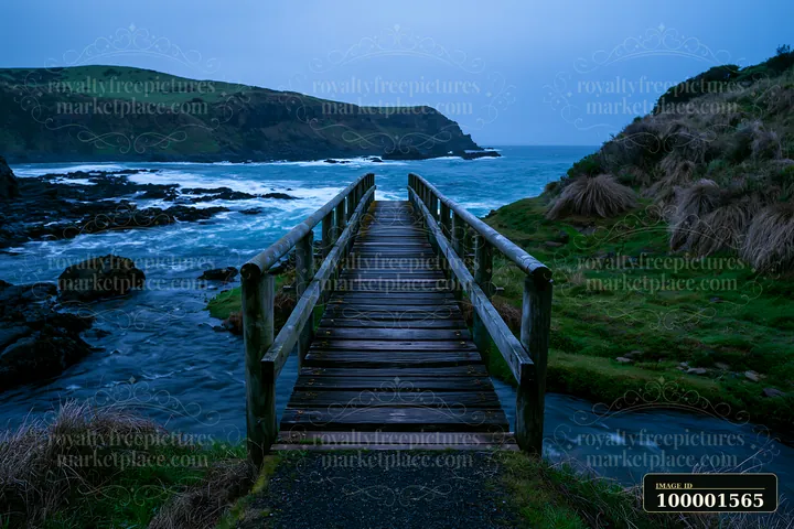 A wooden bridge stretches over water, leading to a rugged coastline beneath dramatic cliffs.
