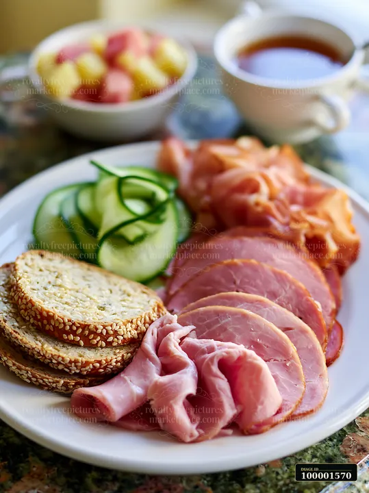 A breakfast plate featuring cold cuts, cucumber slices, and sesame seed bread is served alongside a bowl of fresh fruit and a cup of tea.