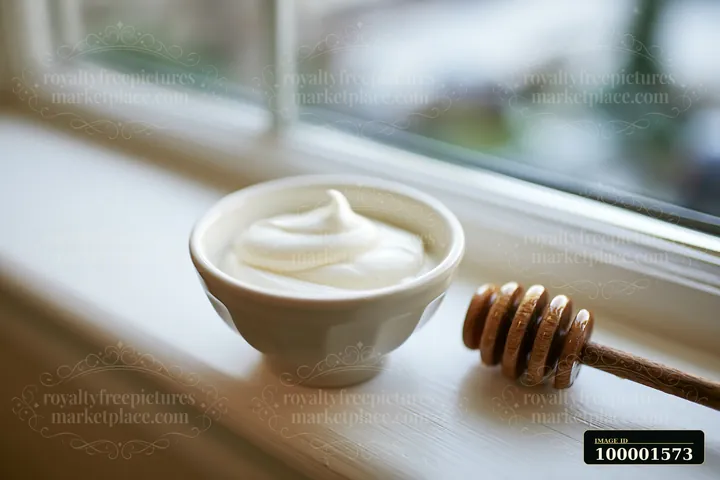 A bowl of cream sits on a windowsill next to a honey dipper, creating a simple kitchen scene.