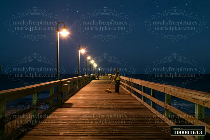A worker sweeps the wooden boardwalk under bright lights while a starry night sky hangs above the ocean.