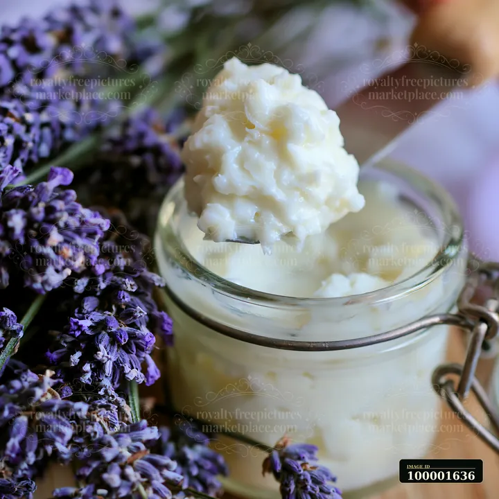 A spatula holds a creamy substance above a glass container filled with cream, set against a backdrop of lavender flowers.