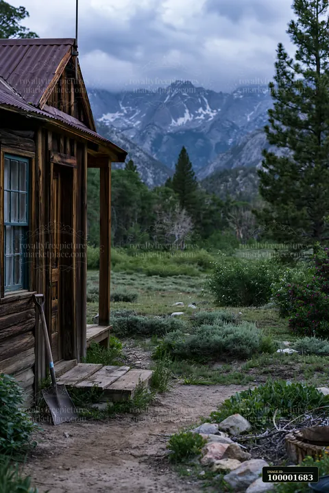 A rustic cabin stands at the edge of a meadow, with dramatic clouds around alpine peaks in the background.
