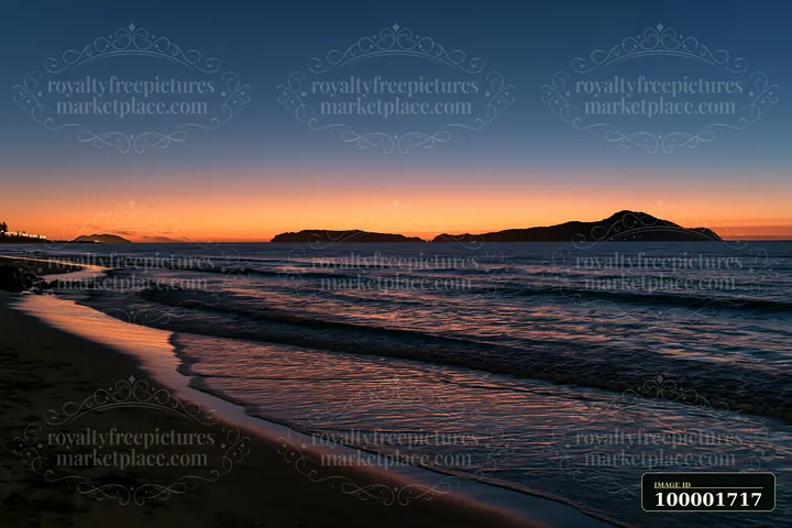 Waves gently lap at the sandy beach as the sun sets behind an offshore island, creating a colorful horizon.