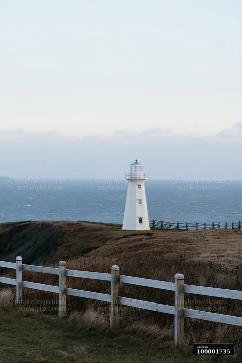 A lighthouse stands on a grassy cliff by the sea with a wooden fence in the foreground.