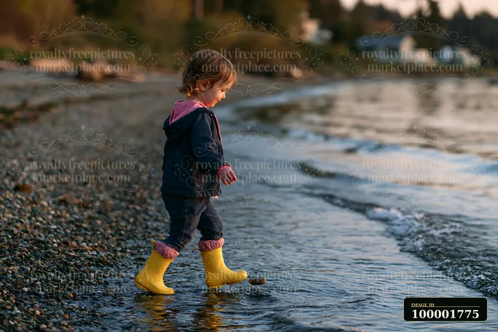A child in bright yellow boots walks along a pebbly coastline beach during sunset, exploring the shore where the water meets the land, surrounded by trees and d