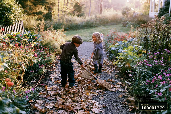 Two children play in a farmhouse garden, sweeping fallen leaves into piles on a path lined with flowers.
