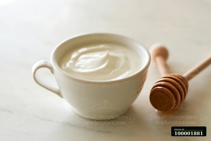 A cup filled with cream sits next to a wooden honey dipper on a light surface.