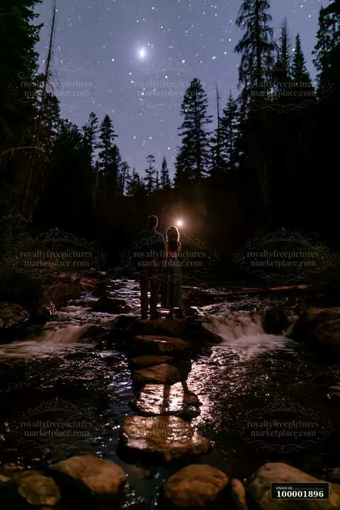 A couple stands together on rocks in a stream, surrounded by a forest at night.