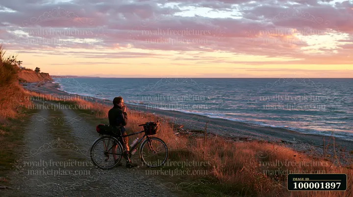 A person stands beside a bicycle on a dirt path, watching the sunset over the pebbly coastline beach.