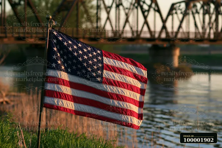 An American flag flying in strong wind stands by a riverbank with a bridge in the background.