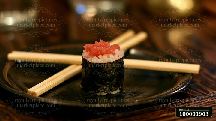 A spicy tuna roll piece sits on a black plate beside wooden chopsticks.