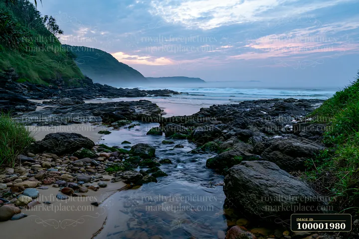 The coastline with exposed rocks features a stream flowing through the area, surrounded by grass and sand as waves lap gently at the shore during twilight.