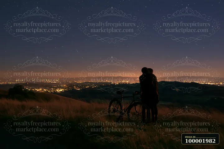 A couple kisses while standing on a hillside, wrapped in a blanket beneath a starry sky.