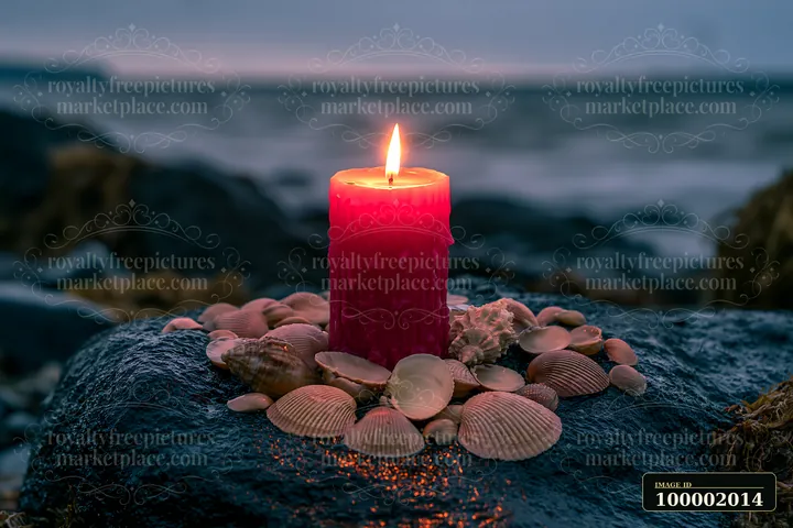 A red candle burns in the center of seashells arranged on a rock by the ocean.