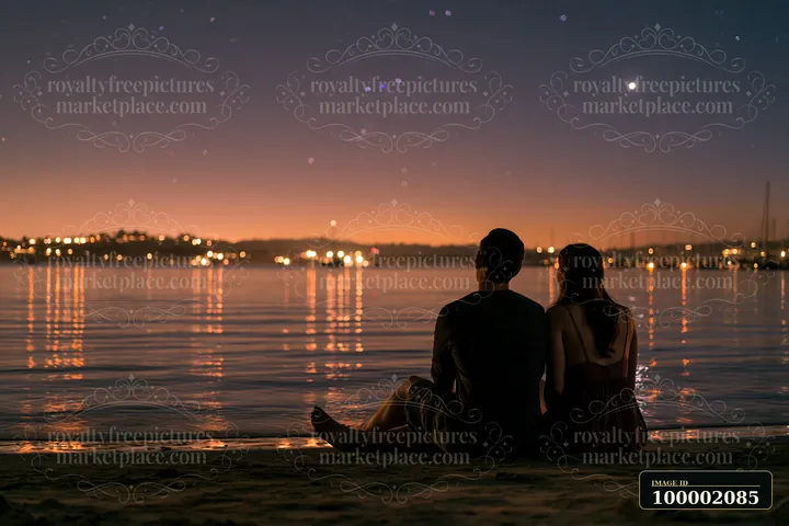 A couple on a beach under stars sits close together, looking at the night sky.