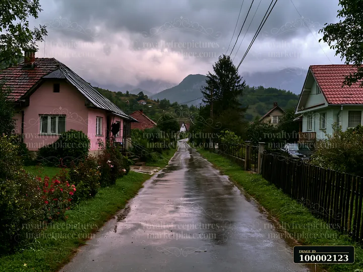 Dark clouds gather over the mountain range as rain falls on a quiet rural road lined with houses.