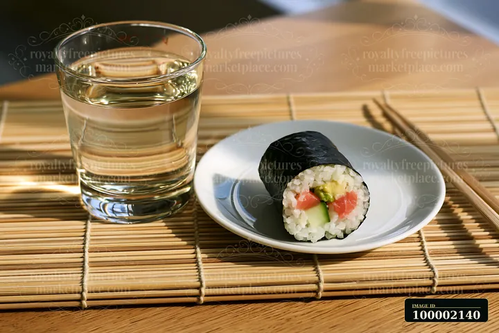 A single sushi roll on bamboo mat is placed next to a glass of water.