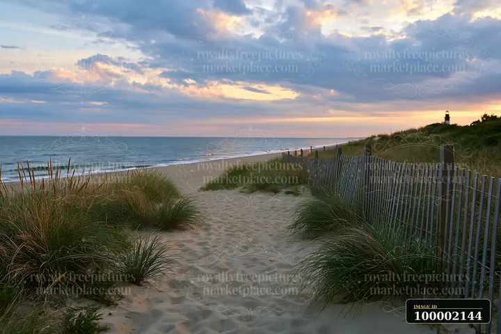 A lighthouse framed by coastal dunes stands near the shore as the sun sets, casting light on the water and clouds.