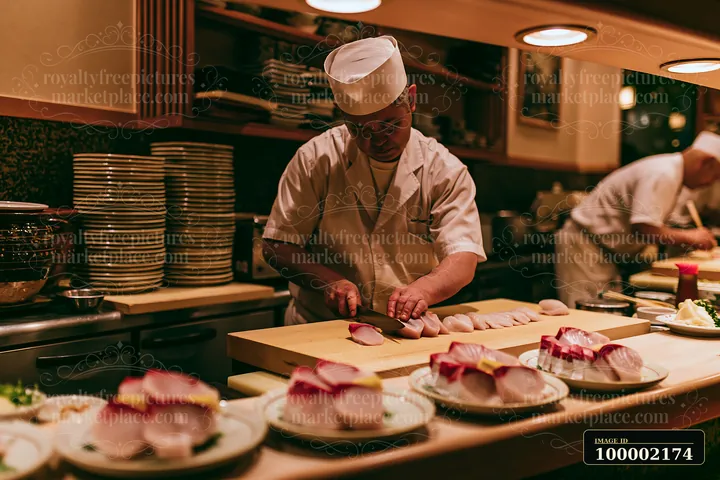 A chef is slicing sushi at a counter in a restaurant kitchen, with neatly arranged plates of sushi in front.