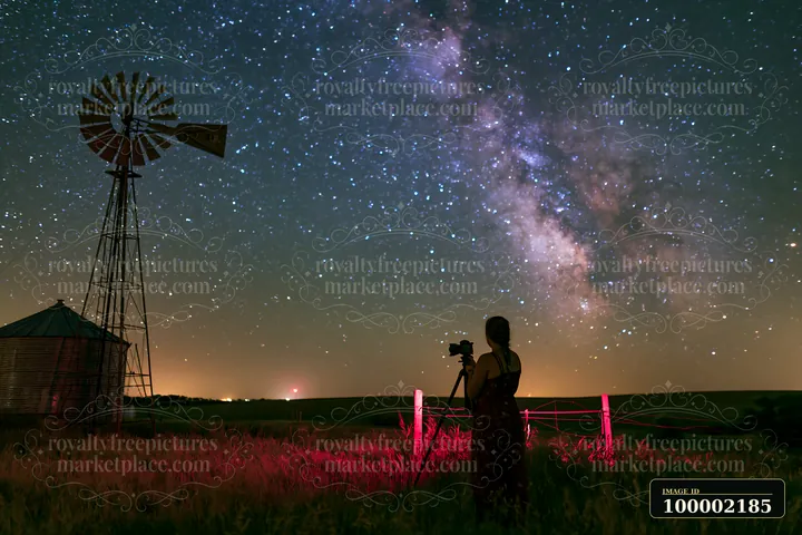 A person using camera under Milky Way stands next to a windmill in a field.