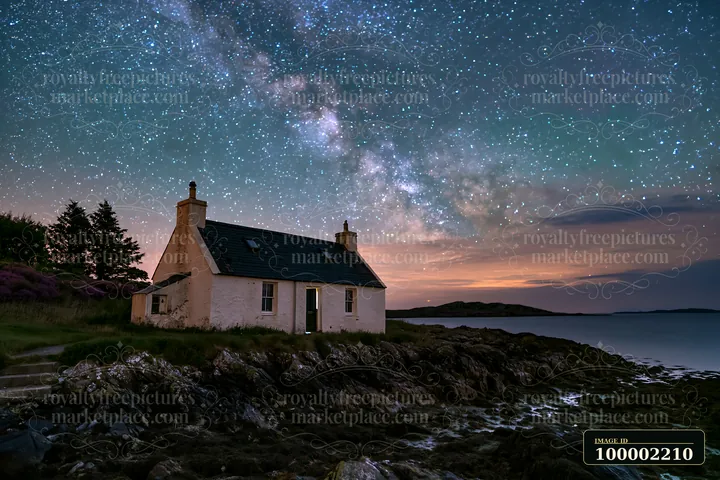 A coastal cottage stands on rocky shores while stars fill the night sky above.