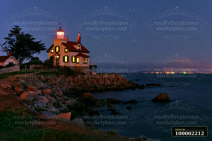 A lighthouse house under stars stands by the water with lights on in the windows.