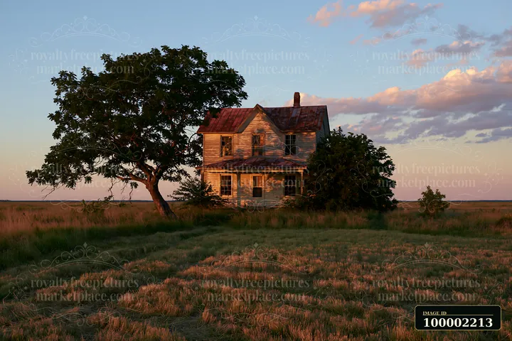 A rustic farmhouse stands under a large tree as the sun sets in a wide-open field.