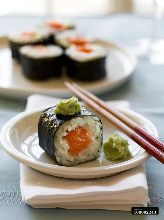 One salmon maki piece with wasabi sits on a white plate, alongside red chopsticks and a napkin.
