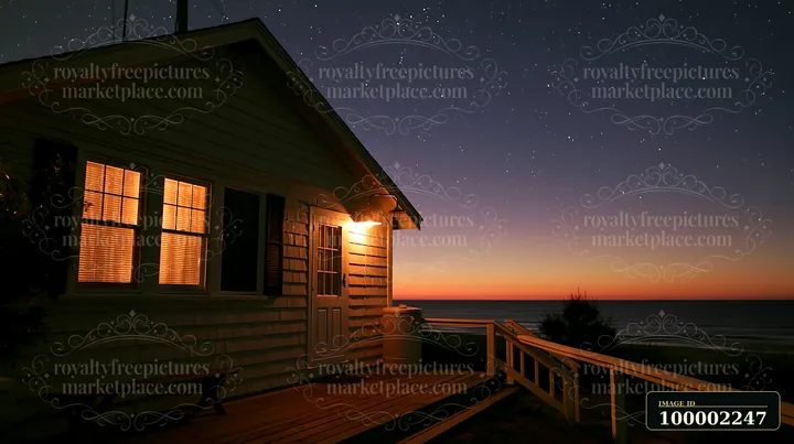 A wooden coastal home sits by the ocean, illuminated by warm lights at night.