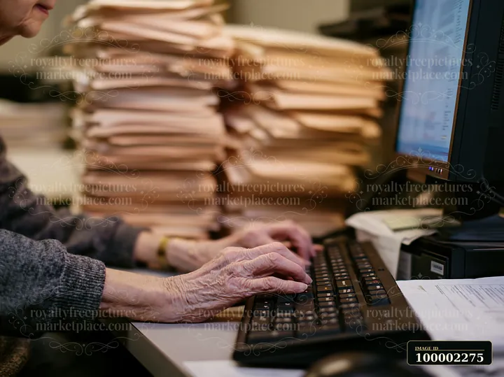 A senior woman is typing at a computer with shaky hands, surrounded by stacks of papers on her desk.