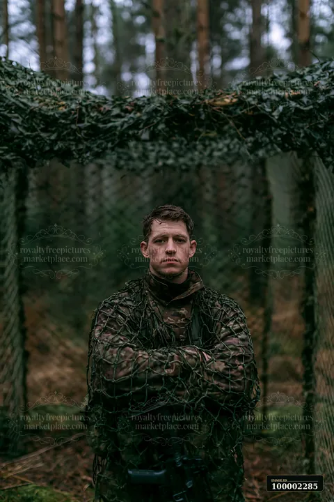 A soldier stands confidently under camouflage netting in a forest.