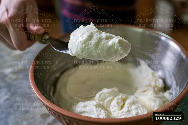 A person holds a spoonful of cream dose with smooth glossy texture above a bowl filled with a thick cream mixture.