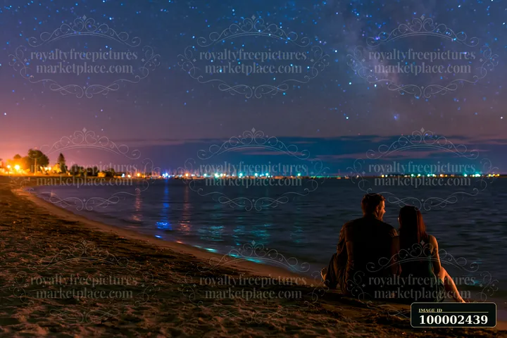 A couple on a beach under stars sits by the water, enjoying the view of the night sky filled with stars.