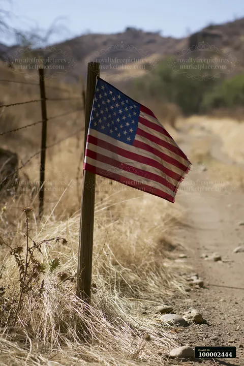 A weathered American flag stands on a post beside a dirt path through dry grass and shrubs.